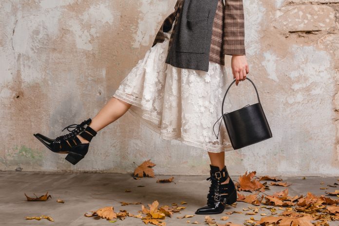 close up legs in boots of stylish woman in jacket coat walking against wall in street, autumn fashion trend, wearing white lace dress, handbag, leaves on the ground, urban style, footwear accessories